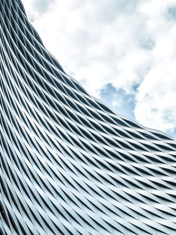 Architecture moderne: Façade abstraite en métal ondulé. Façade abstraite d'un bâtiment moderne; des rubans métalliques ondulés créent un motif de losanges complexes sous un ciel nuageux bleu et blanc.